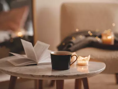 Ein aufgeschlagenes Buch liegt neben einer Tasse und zwei Kerzen auf einem kleinen Tisch.