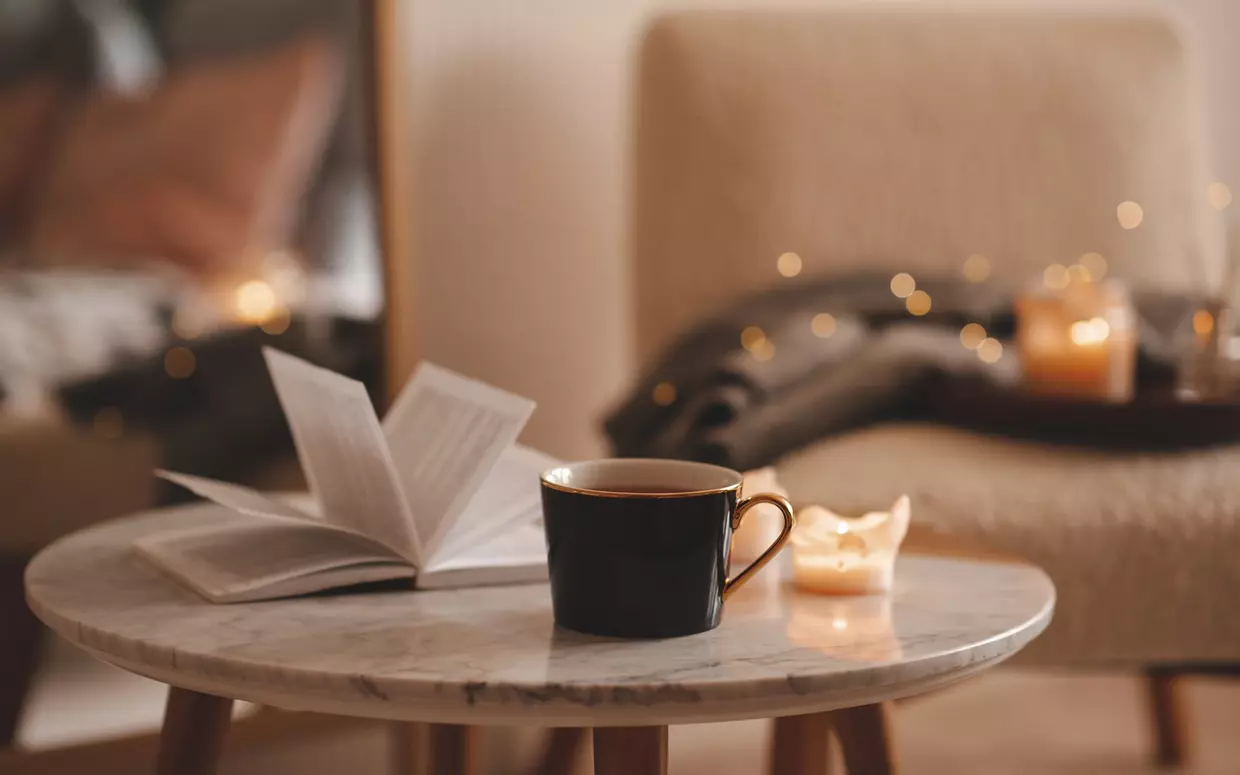 Ein aufgeschlagenes Buch liegt neben einer Tasse und zwei Kerzen auf einem kleinen Tisch.