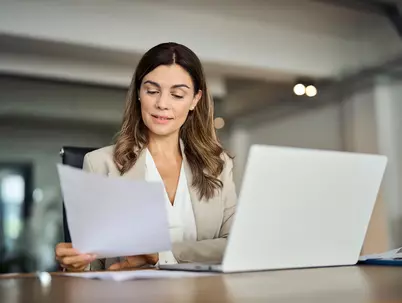 Eine Frau im Business-Outfit sitzt vor dem Laptop und bearbeitet Dokumente.