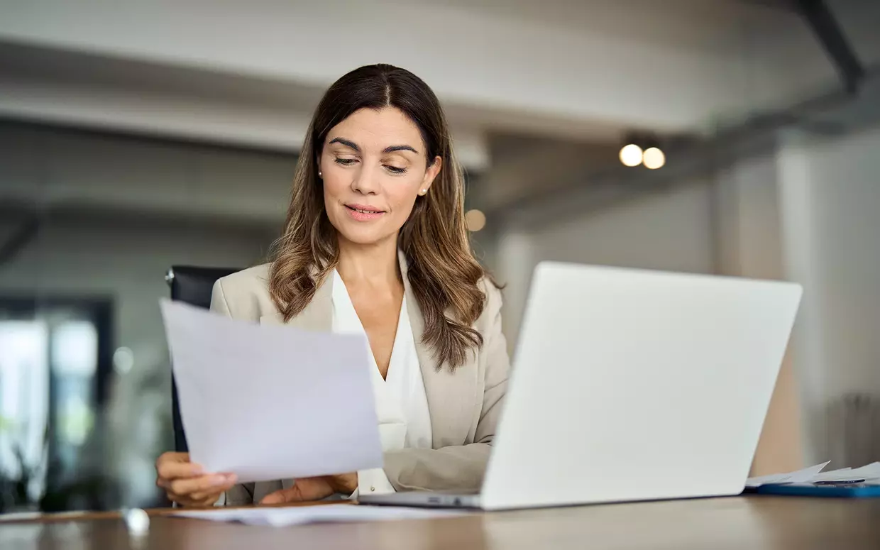 Eine Frau im Business-Outfit sitzt vor dem Laptop und bearbeitet Dokumente.