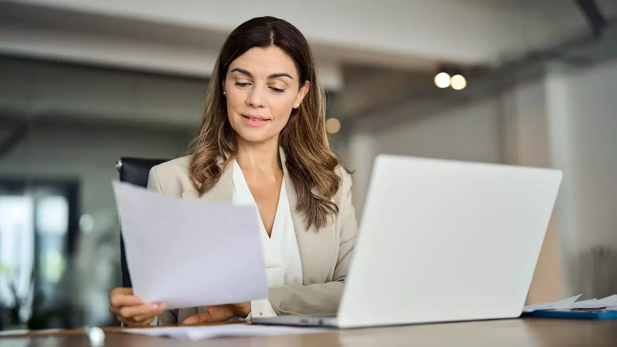 Eine Frau im Business-Outfit sitzt vor dem Laptop und bearbeitet Dokumente.