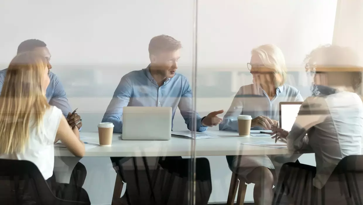 Hinter einer Glaswand halten fünf Personen ein Meeting ab.